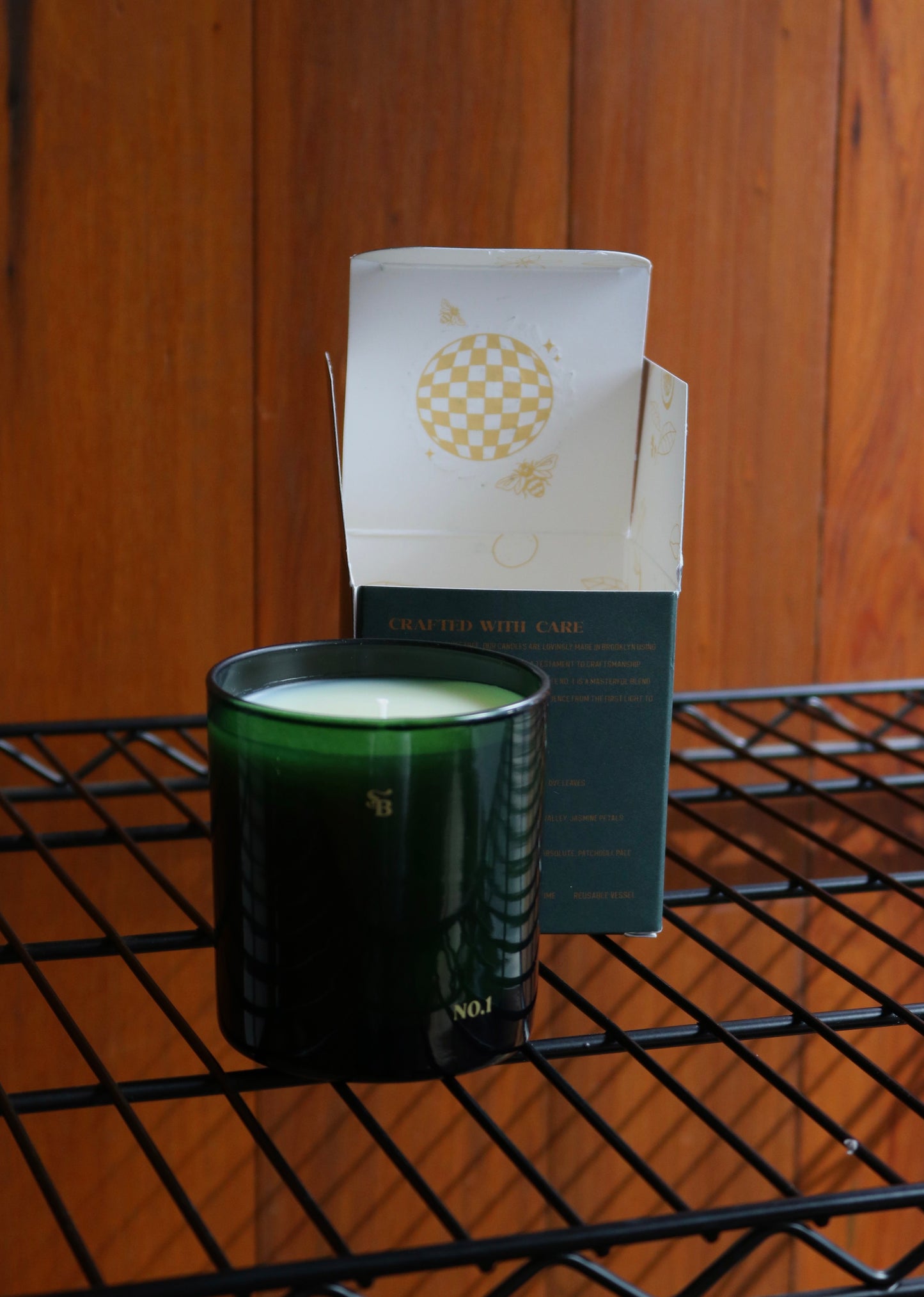 Green candle in a black holder with a white box on a metal shelf against a wooden wall.